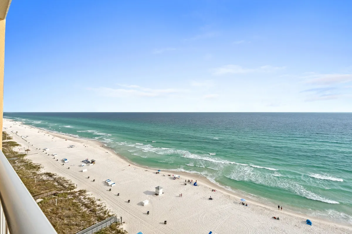 A panoramic view of a lively stretch of white sandy beach and turquoise ocean, dotted with people sunbathing, swimming, playing volleyball, and walking along the shore. The overall vibe is relaxed and energetic, ideal for group activities.