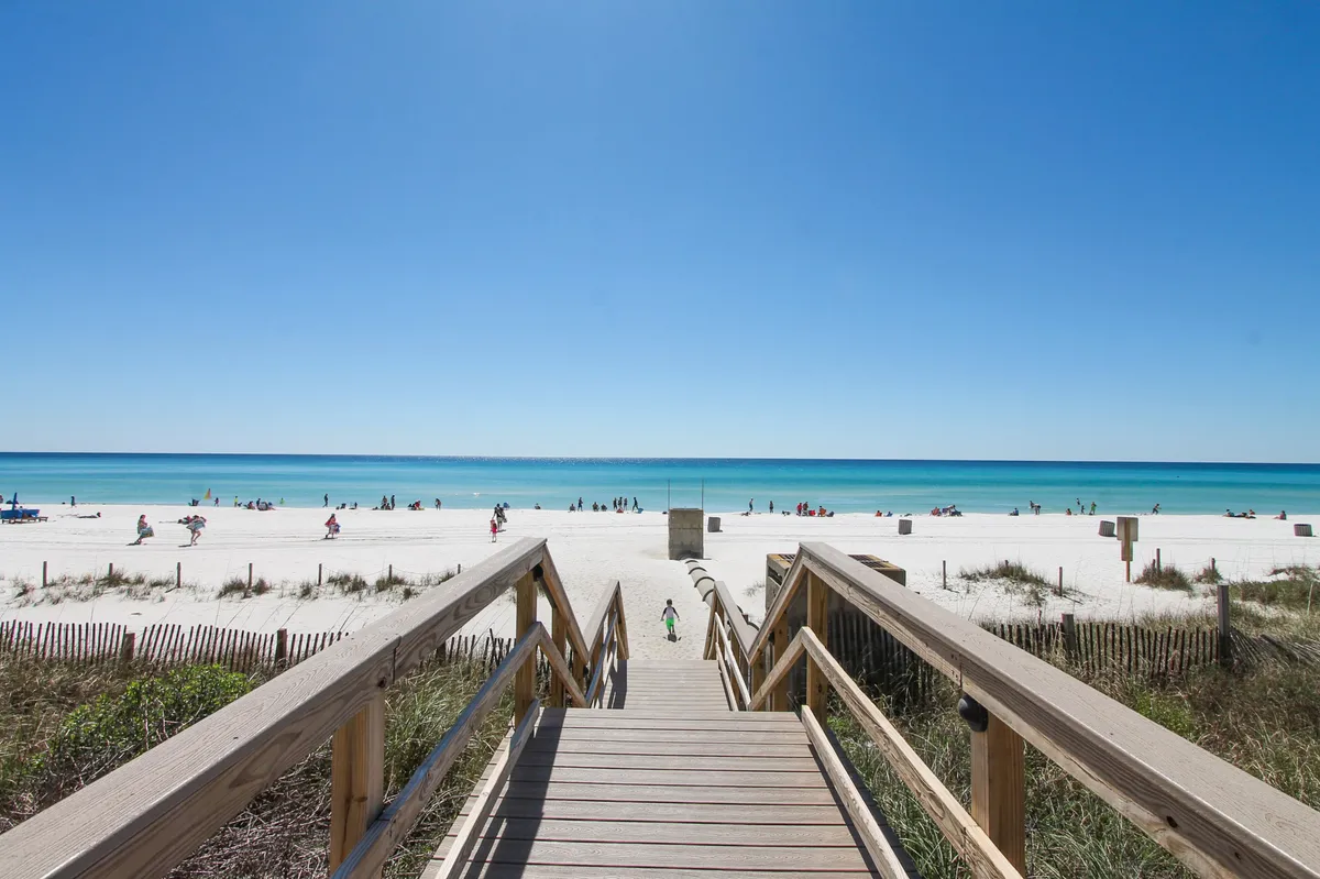 A scenic view of the beach from a wooden walkway, with guests building sandcastles, sunbathing, and playing games on the soft sand. The ocean stretches into the distance, and the mood is relaxed, inviting families to enjoy private beach access.