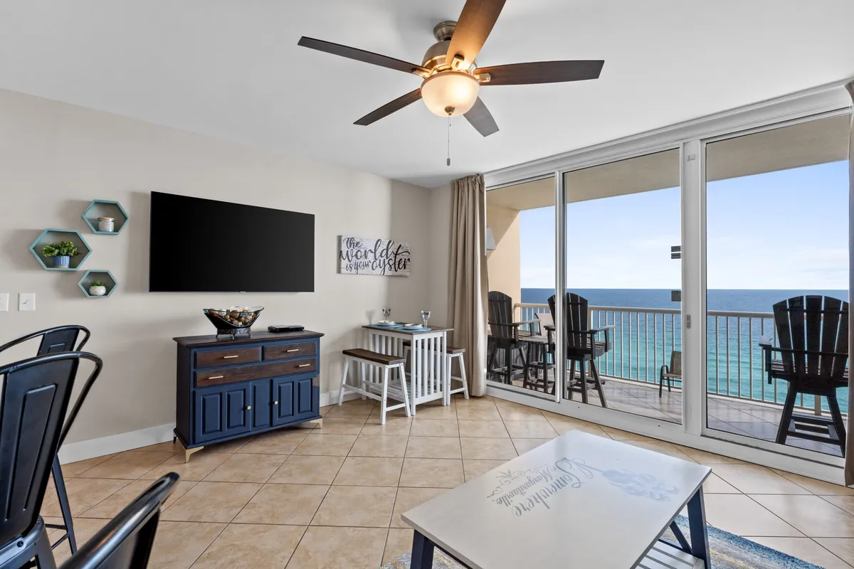 A bright, inviting living room filled with natural light from large floor-to-ceiling windows that offer sweeping ocean views. The stylish coffee table sits in front of a plush sofa, and a modern flat screen TV anchors the entertainment area. The open-plan design flows into a dining space, all oriented toward the view and social interaction.