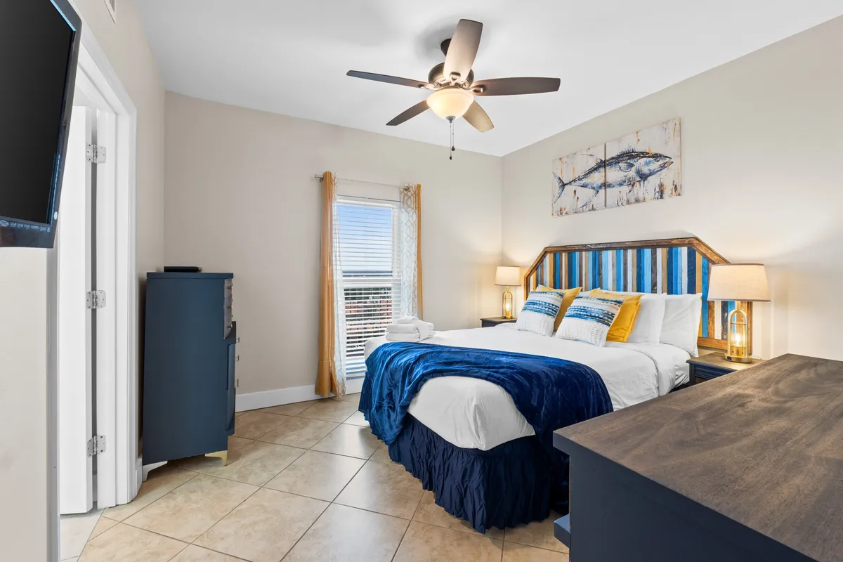 A serene master bedroom featuring a king-sized bed adorned with blue and white bedding, tasteful fish-themed artwork above the bed, and bedside tables with lamps. The room receives soft natural light, creating a tranquil and restful environment.