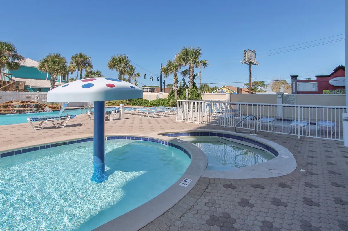 An exterior family pool area with a whimsical mushroom umbrella fountain, a large swimming pool, lounge chairs, and palm trees. Children can be imagined playing under the fountain, with families gathered for a relaxing day together.