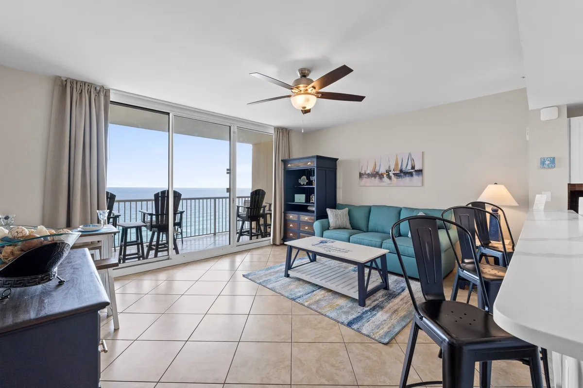 A sun-drenched living room with a teal sofa and nautical-themed cushions faces large sliding glass doors that lead to a private balcony with panoramic Gulf views. A navy blue cabinet and modern ceiling fan complete the space, and the inviting seating arrangement is ideal for socializing or relaxing.
