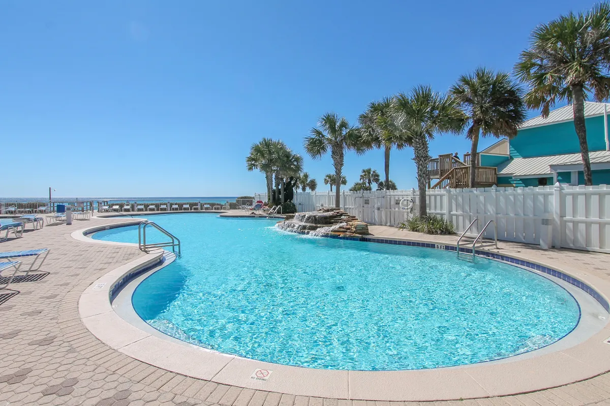 A large, curved swimming pool with a striking waterfall feature is surrounded by palm trees, with the ocean visible in the background. Poolside lounge chairs are arranged along the sunny deck, and the pool area is spacious and inviting.