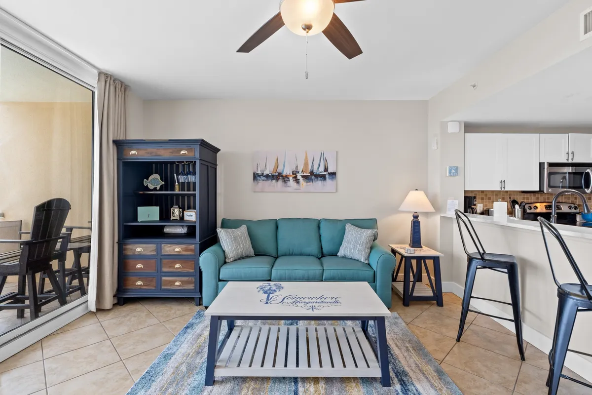A welcoming living area with a teal sofa and accent pillows, a blue cabinet with decorative items, a coffee table with a nautical design, and a kitchen bar with stools, all set in a relaxed, coastal-themed space.