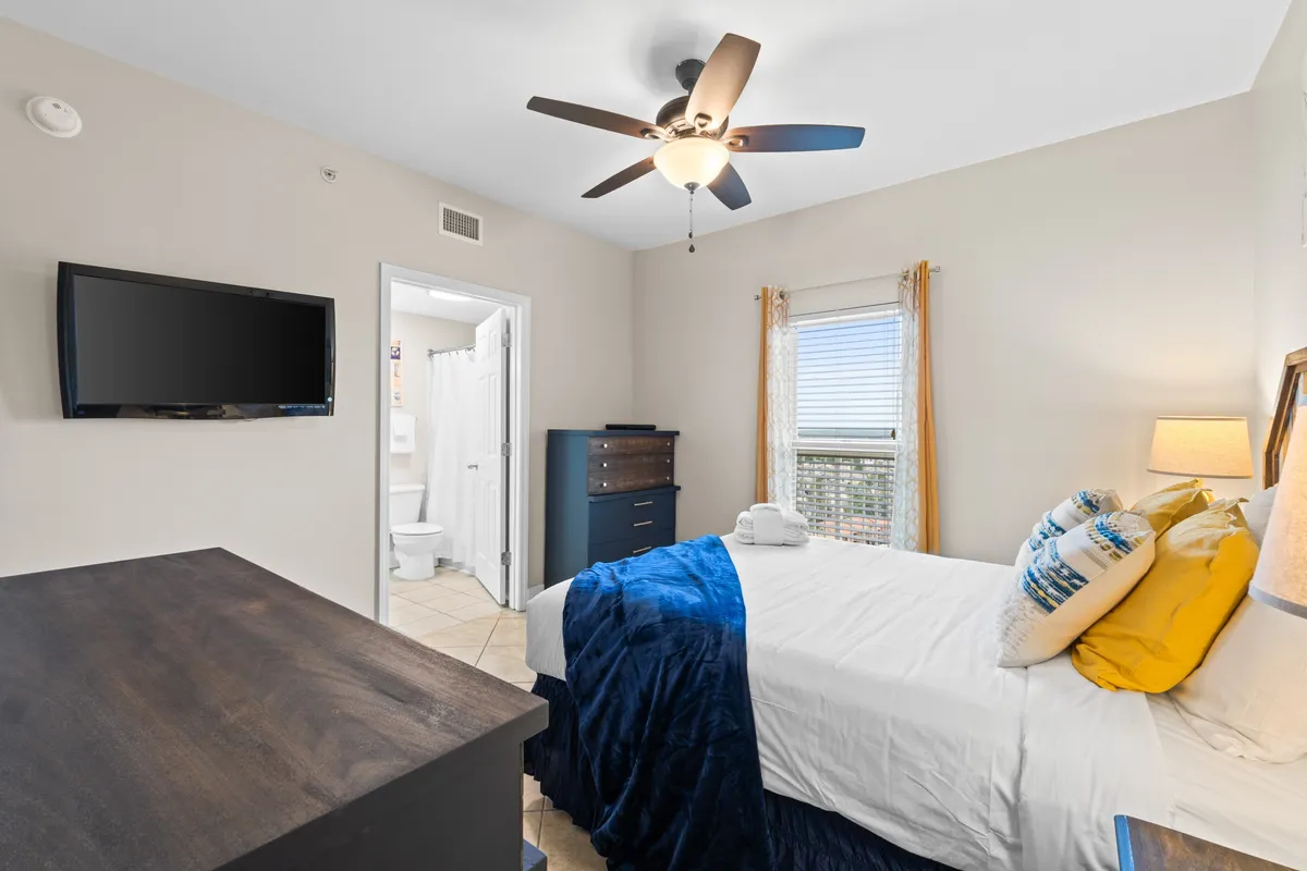 A cozy second bedroom featuring a comfortable queen-sized bed with bright pillows and a navy throw blanket, a wall-mounted flat screen TV, a window letting in natural light, and an open doorway to a private bathroom.