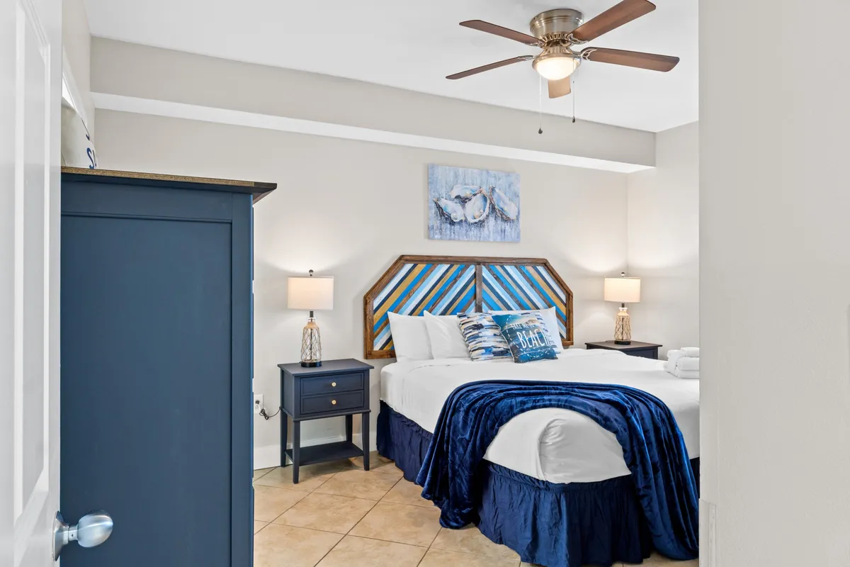 A serene master bedroom with a king size bed adorned with decorative pillows, matching nightstands with lamps, a ceiling fan with light, and nautical-themed wall art, all bathed in soft natural light.