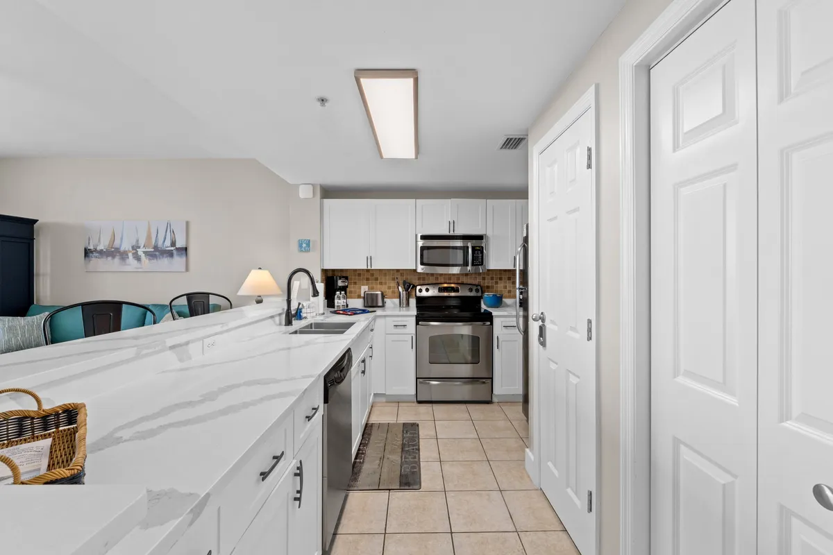 A long white marble countertop runs the length of the kitchen, accompanied by stainless steel appliances and white cabinetry. A decorative backsplash adds coastal flair, and the overall space is open and inviting, perfect for cooking and gathering.