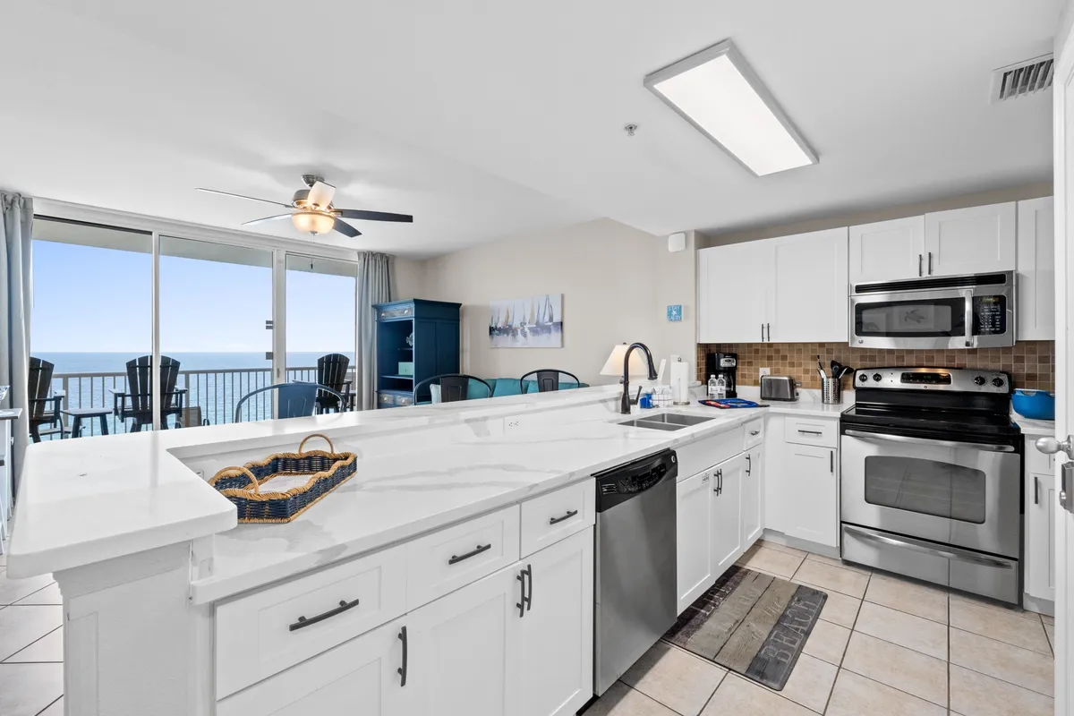 An open-plan kitchen and dining area with a large white marble countertop island, blue cabinetry, and modern appliances. Floor-to-ceiling sliding glass doors open to a private balcony with stunning ocean views, while the adjacent dining area invites group meals.