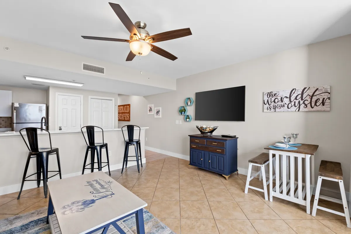 A cozy, open-concept living space with a wall-mounted flat screen TV, blue cabinet with decorative bowl, kitchen counter with three black bar stools, and a small dining table with bench seating. The space feels welcoming and perfect for socializing or relaxing after a day of resort activities.