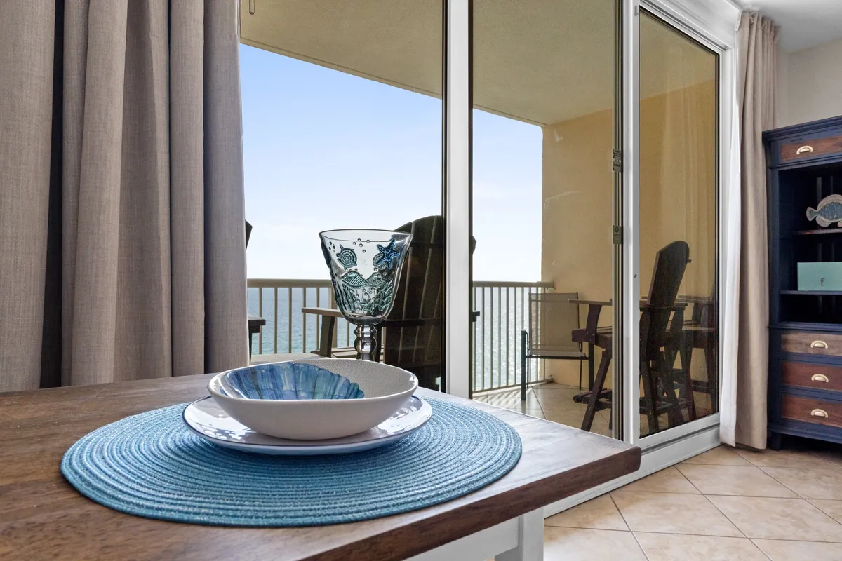 A dining table set with blue-themed dishware sits next to a glass door leading out to a balcony with ocean views. Balcony furniture is visible, and a blue cabinet with decorative items adds a pop of color to the serene, coastal-inspired dining area.