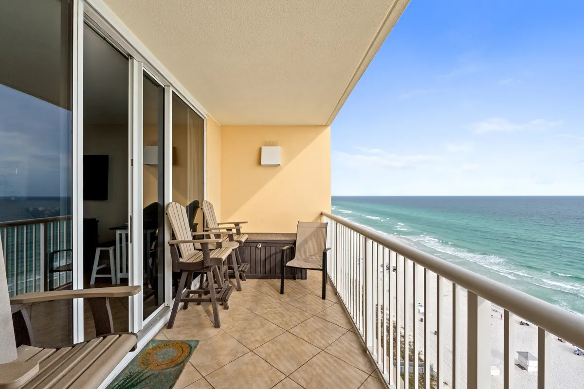 A private balcony set with comfortable seating overlooks an expansive ocean and white sandy beach. The clear blue sky and horizon create a serene coastal backdrop, making it a perfect spot for relaxing or sharing moments with family and friends.