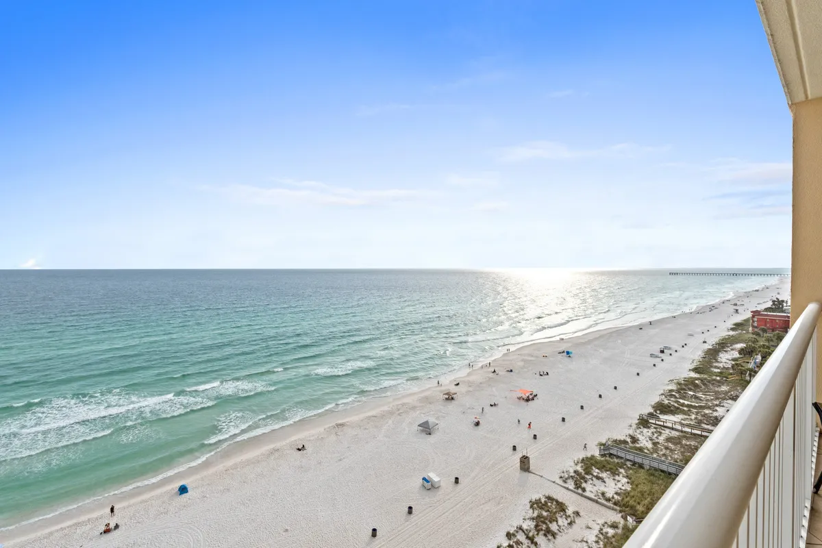 This image captures a private balcony overlooking the sparkling turquoise waters of the Gulf of Mexico, with a white railing framing the view. The wide sandy beach below is dotted with beachgoers and umbrellas, and a distant pier extends into the water. The mood is calm and rejuvenating, evoking the perfect spot for peaceful morning coffees or romantic sunset dinners.