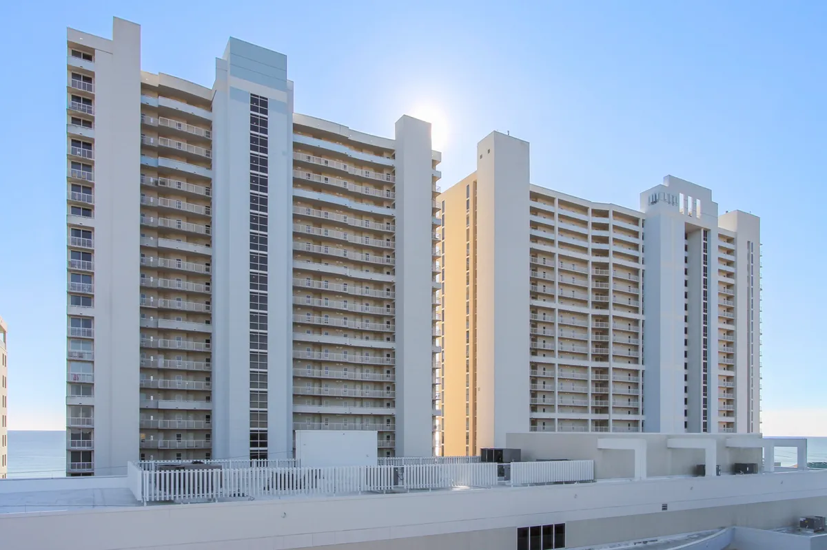 A multi-story resort building with balconies faces the ocean, set against a clear bright sky. The scene shows the inviting beachfront location, resort amenities, and easy access to the beach. The ocean is visible in the background, underscoring the proximity to the water and the overall vacation atmosphere.