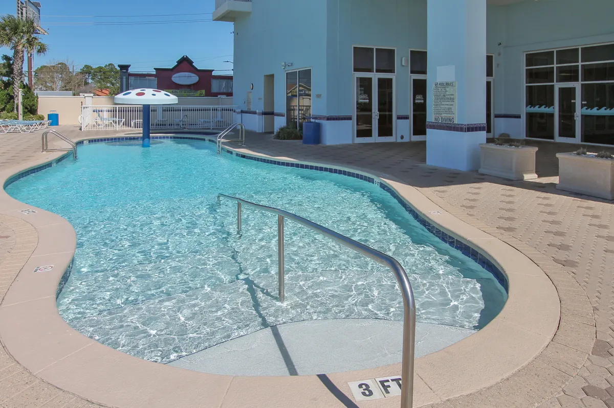 The exterior pool area features a curved swimming pool with a blue mushroom water feature, poolside lounge chairs, and a modern building facade. Children can play under the water feature while adults socialize or relax nearby.