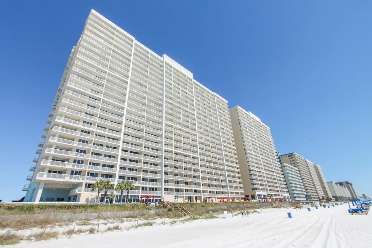 This image showcases the Majestic Beach Resort high-rise building with private balconies, white sandy beach in the foreground, and a clear blue sky overhead. The proximity of the resort to the beach is evident, as well as the inviting resort environment.