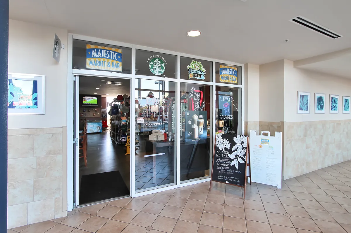 A bright, inviting interior of the onsite market, featuring glass entrance doors with signage, a chalkboard menu sign, product displays, and colorful tiled floors and wall decor. The atmosphere is welcoming and casual, ideal for grabbing snacks or essentials.
