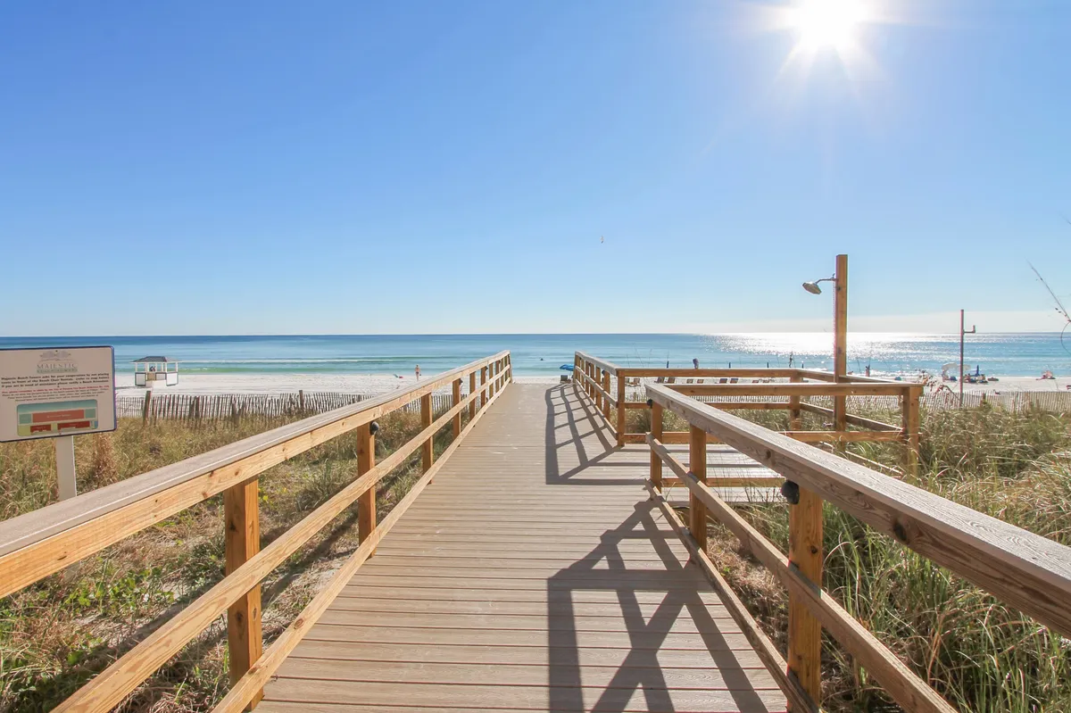This image presents a classic boardwalk leading directly to the calm ocean waters, flanked by beach fencing and dune grasses. The bright sun and clear sky evoke an inviting, carefree beach day, with a sense of anticipation for water activities and relaxation.