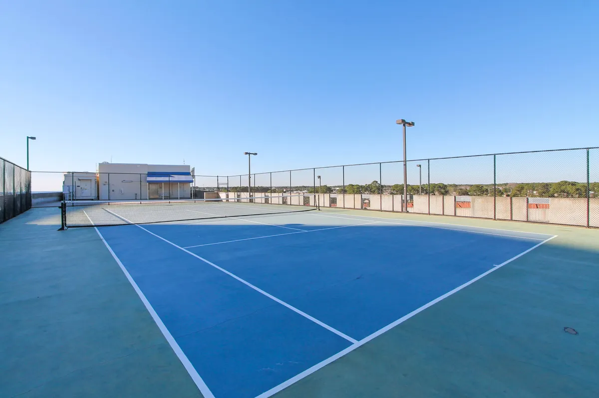 A tennis court with net and safety fencing is shown under a clear blue sky, with the resort building structure visible in the background. The court invites guests to engage in friendly matches or light exercise steps from their accommodation.