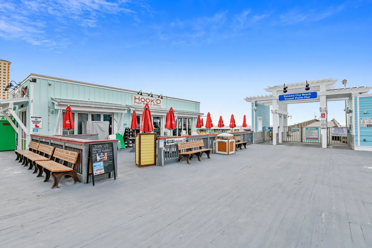 This image features the Hook'd Beach Bar & Grill with its distinctive red umbrellas, benches lining the Russell Fields Pier entrance, and a clear view of the ocean under a blue sky. The scene captures people dining, socializing, and relaxing by the beach, with easy access to the pier and lively coastal atmosphere.