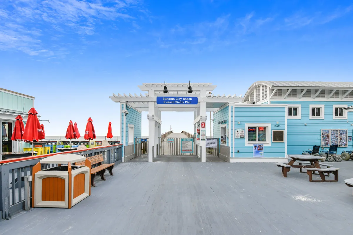 The entrance to the Panama City Beach Russell Fields Pier is shown with vibrant red umbrellas, a light blue building, benches, and picnic tables. The scene is lively and welcoming, suggesting easy access to beachfront dining, socializing, and sunset strolls just steps from the resort.