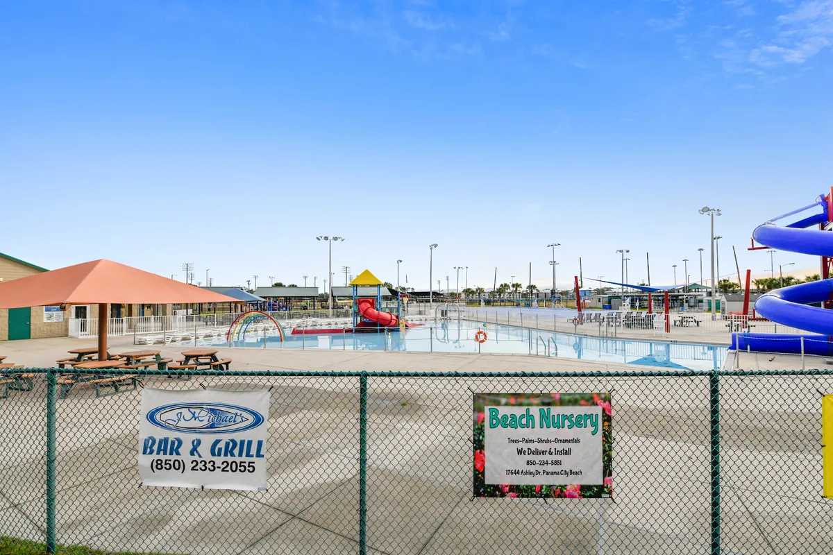 This lively exterior scene spotlights a children's splash pad with colorful water features and slides, a large blue spiral water slide, shaded picnic tables, and a fenced, safe perimeter. Families are seen enjoying the area, emphasizing the kid-friendly amenities.